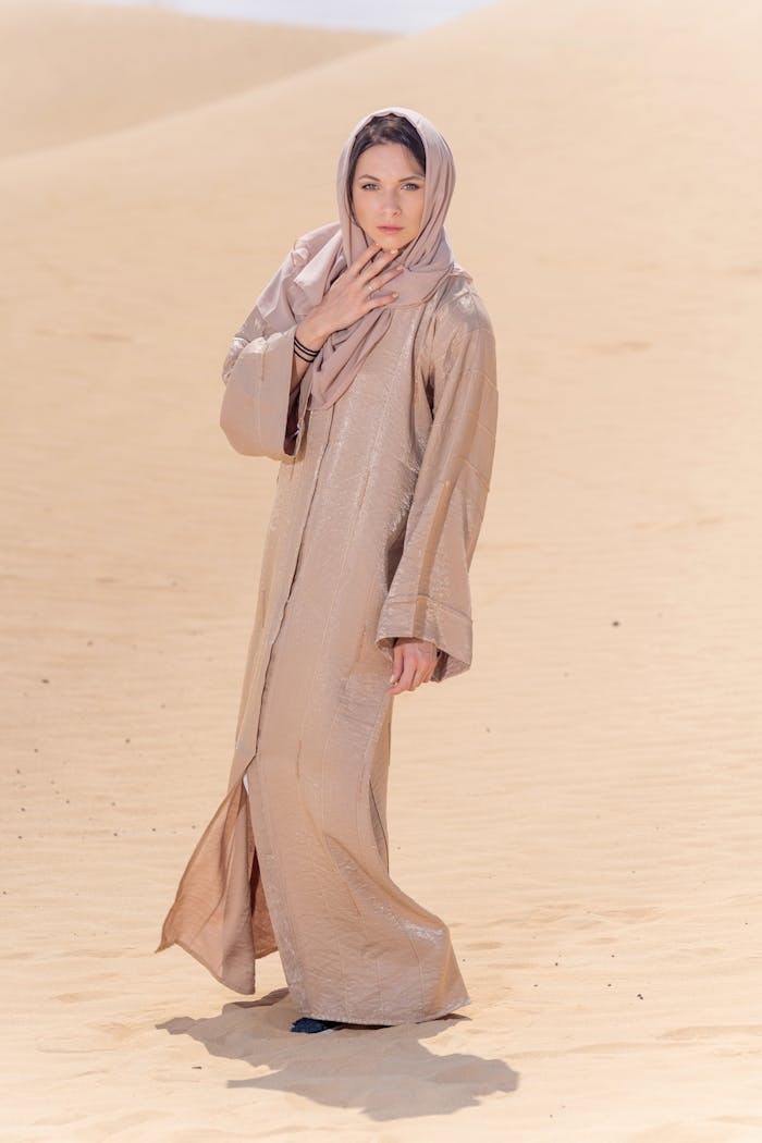 A woman wearing a beige abaya and headscarf stands gracefully in the Dubai desert sands.